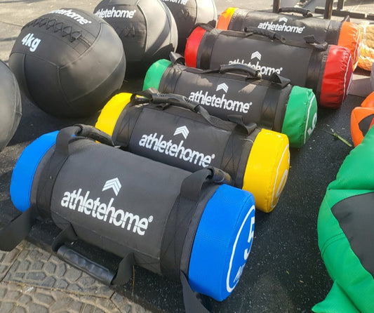 A collection of black weighted training bags with different colored handles, labeled 'ATHLETENHOME', displayed outdoors with a logo and equipment in the background.